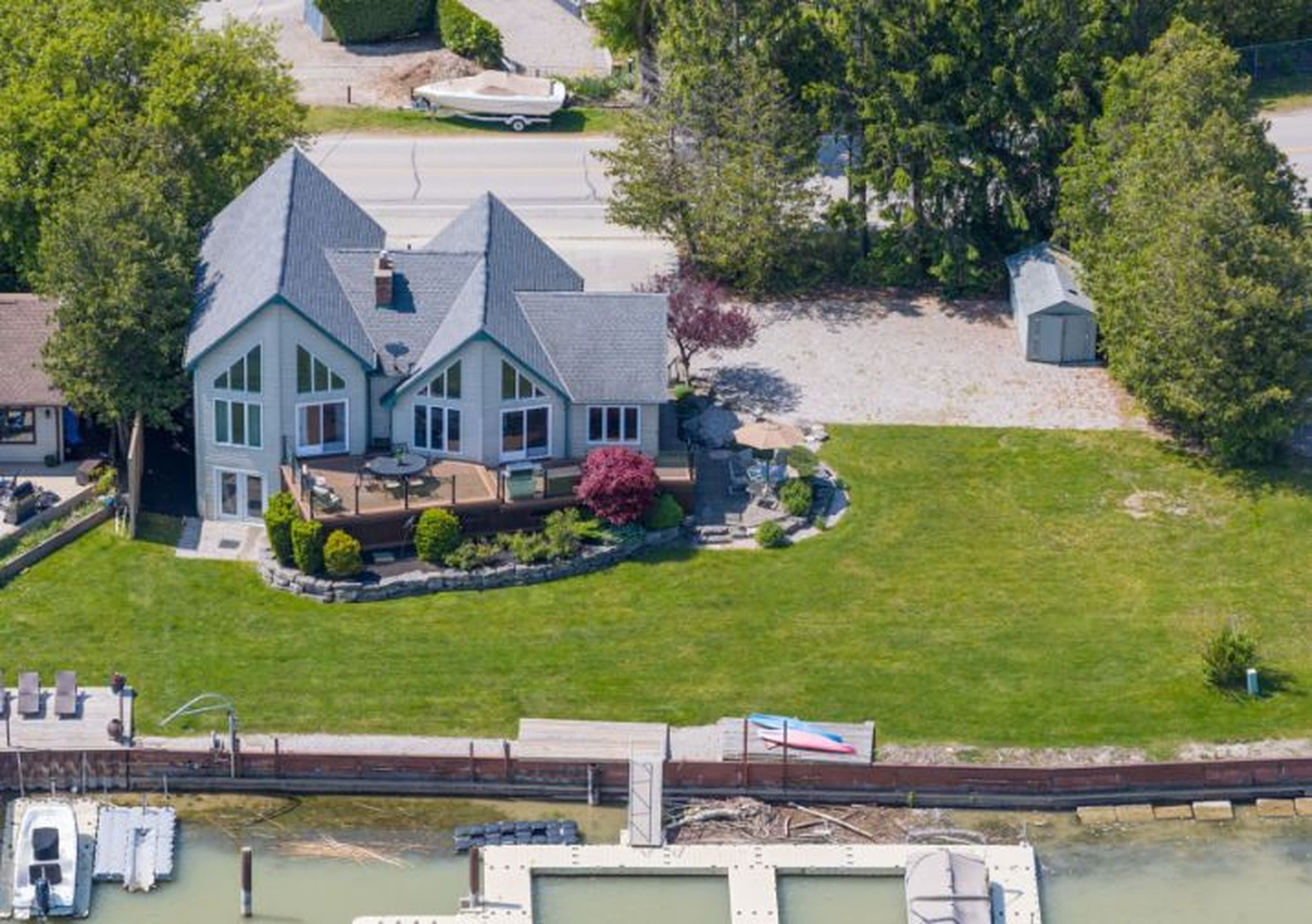 Aerial view of a grey waterfront house with a large deck, green lawn, and dock.