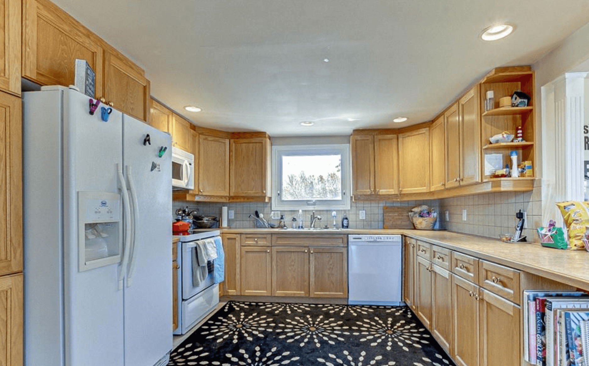 Kitchen with light wood cabinets, white appliances, a window, and a black patterned rug.