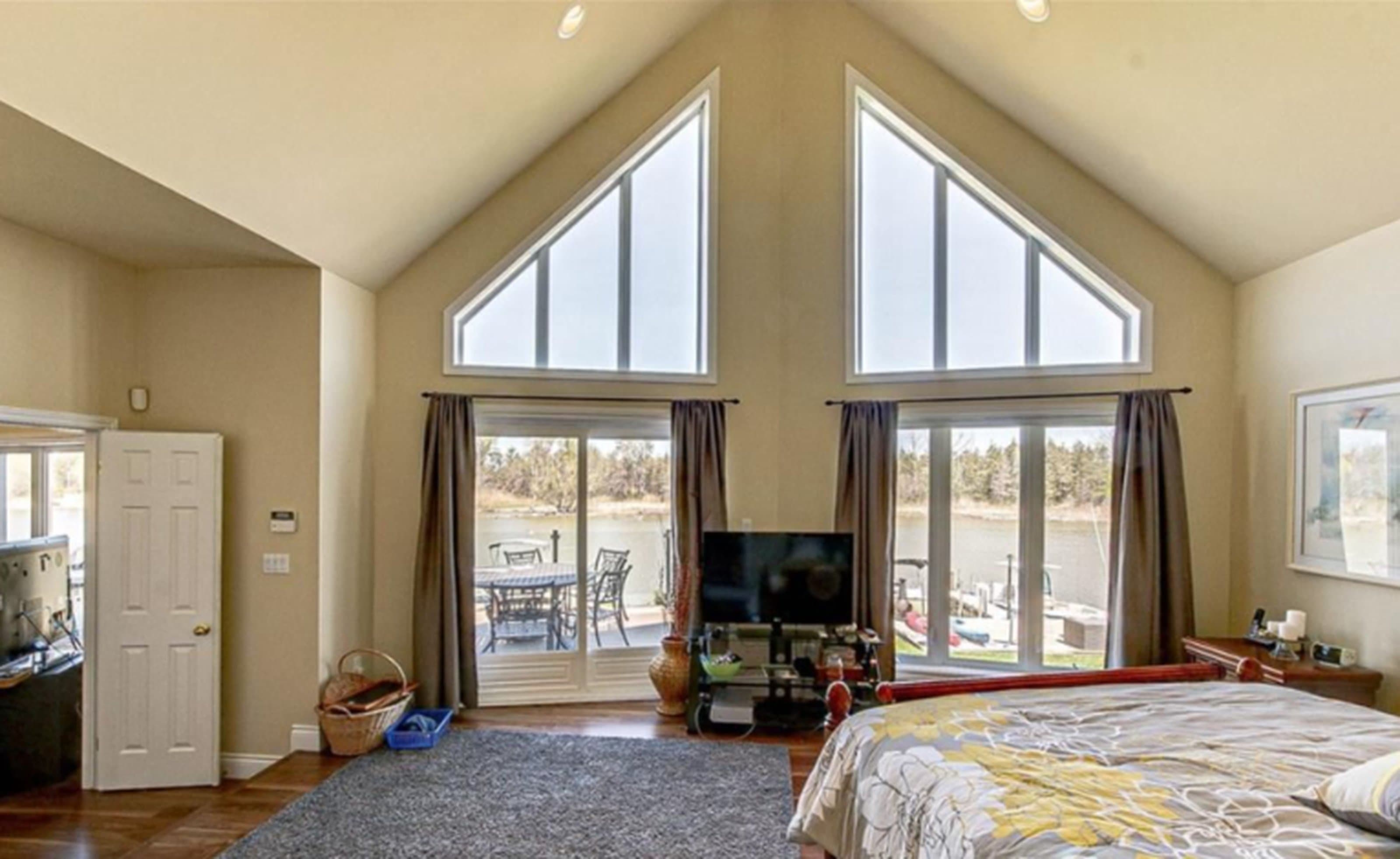 Bedroom with vaulted ceilings, large triangular windows, and glass doors overlooking a scenic lake view.